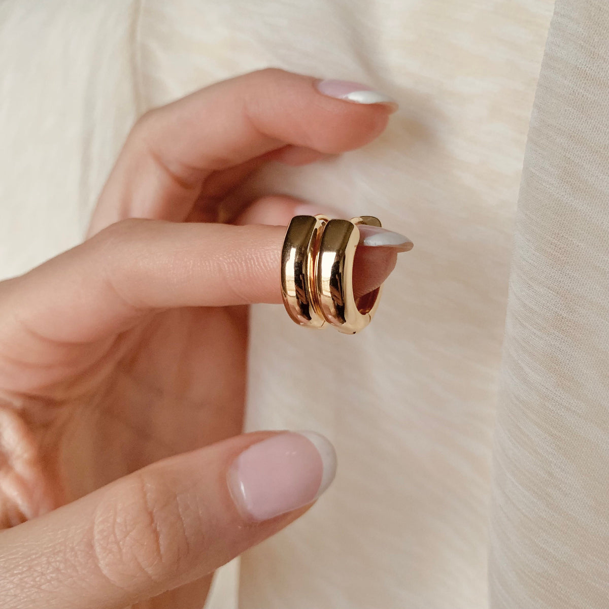 A close-up photo of a hand holding a pair of 16mm chubbie huggie hoop earrings, which are gold-plated and circular in shape. The background features a soft, beige fabric, adding a neutral and elegant setting to the image.