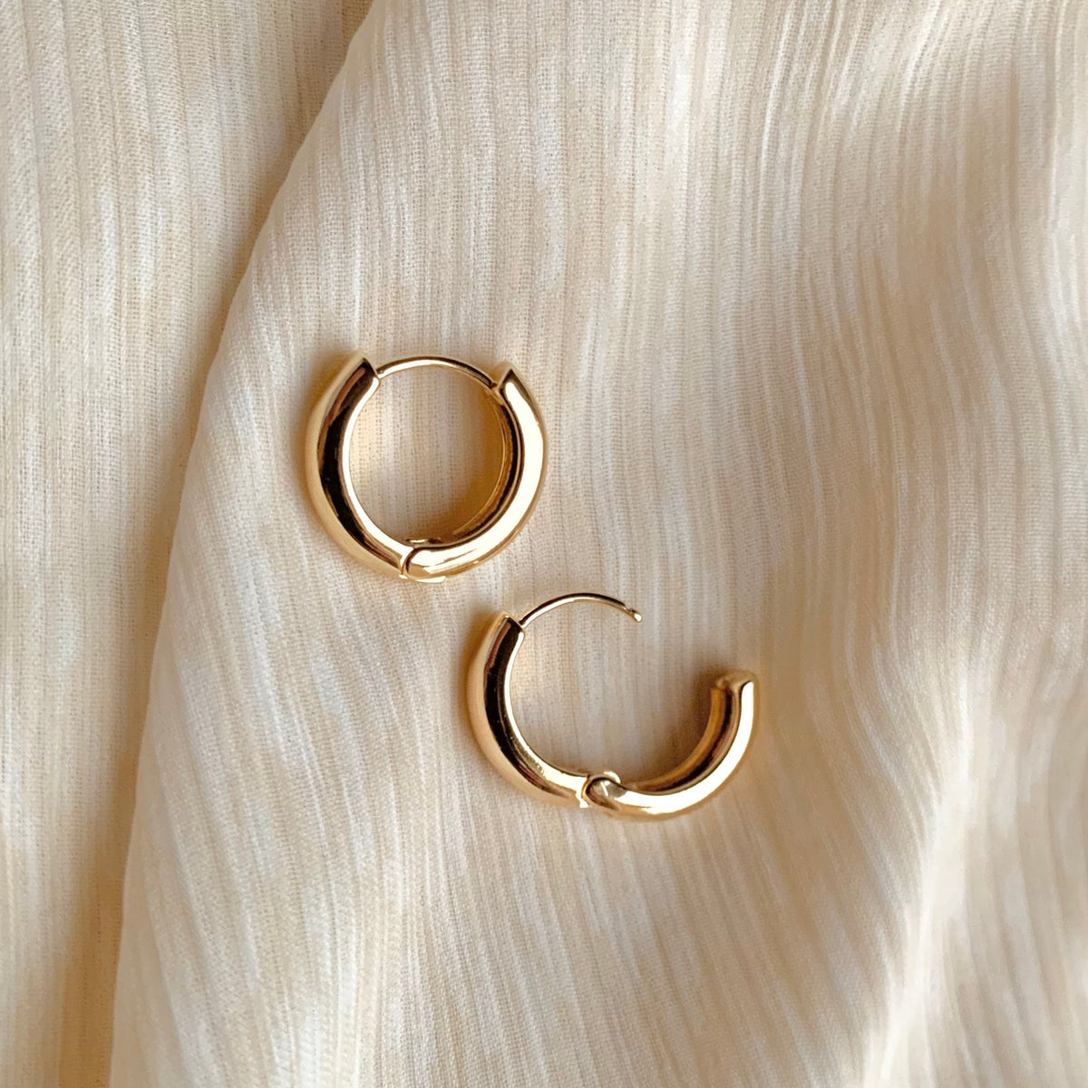 Pair of 16mm chunky gold huggie hoop earrings with a smooth, gold-plated finish, displayed on soft neutral fabric. Classic, minimalist jewelry perfect for everyday wear.